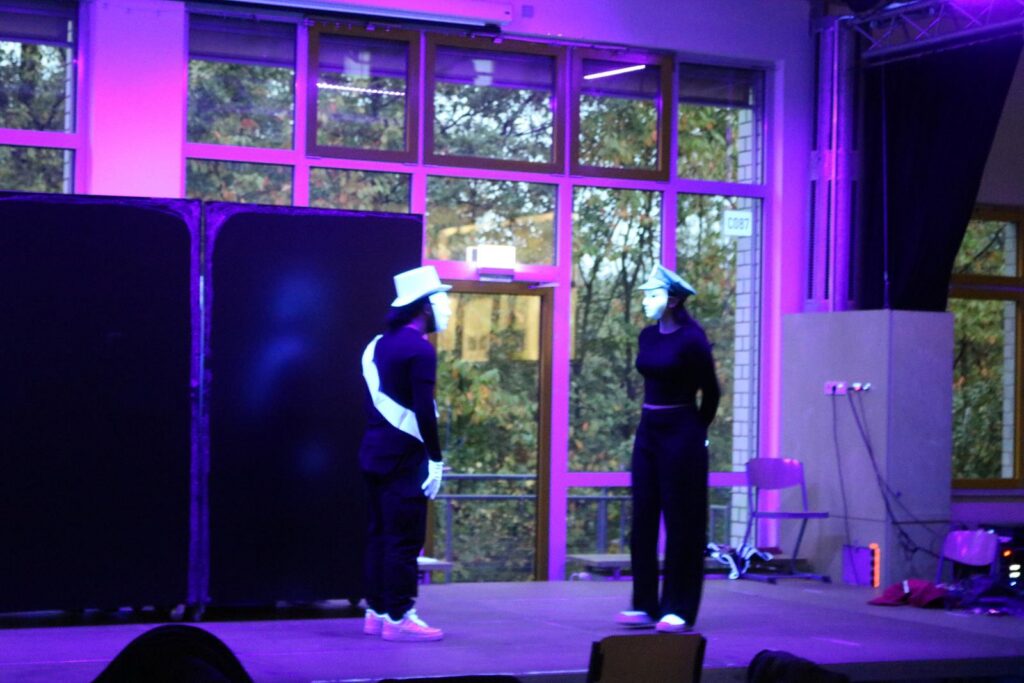 Zwei Personen in Kostümen stehen auf einer Bühne mit buntem Licht.