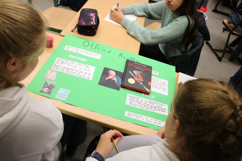 Schülerinnen präsentieren ein Plakat zu einem Thema in einem Klassenzimmer.