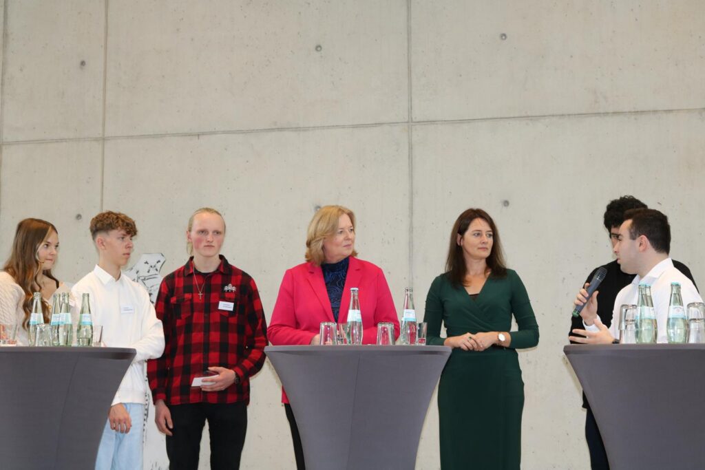 Diskussionsrunde mit mehreren Personen an Stehtischen, Hintergrund ist eine Betonwand.