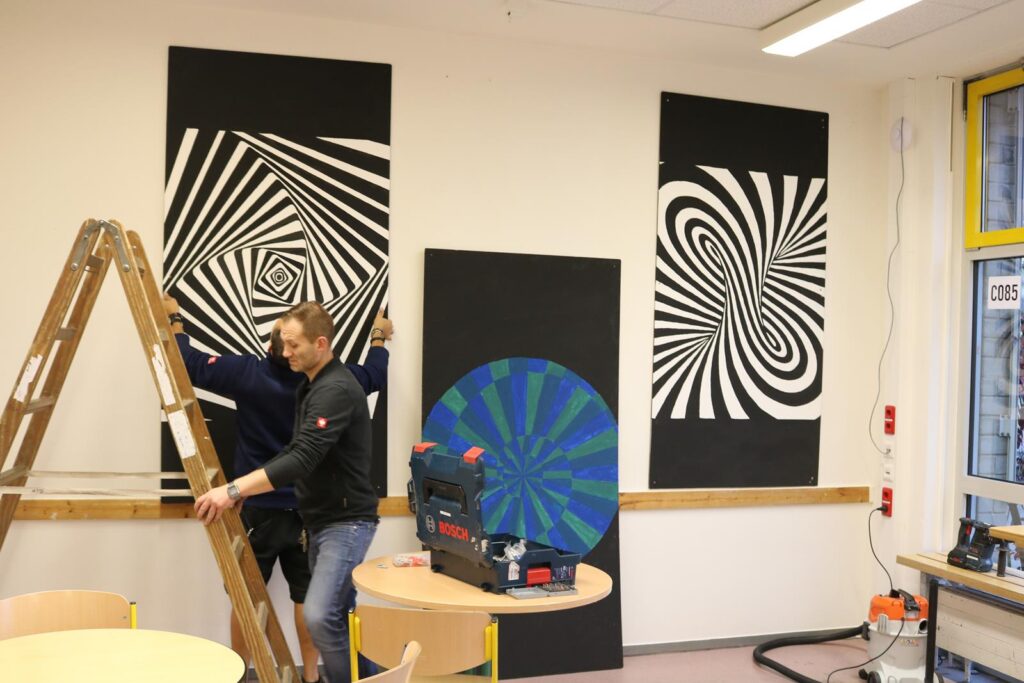 Zwei Personen montieren Kunstwerke an einer Wand, Leiter im Vordergrund.