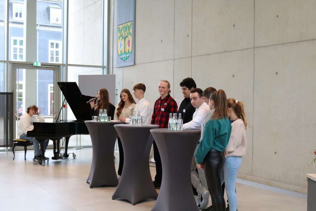 Schüler stehen an Stehtischen, während eine Schülerin Klavier spielt.