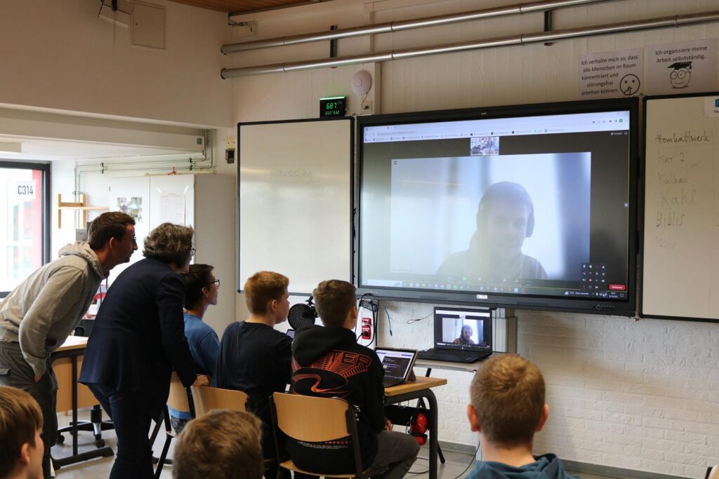 Schüler und Lehrer in einem Klassenraum verfolgen ein Videokonferenz-Meeting auf einem Bildschirm.