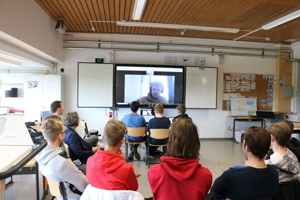 Schüler sitzen im Klassenraum und sehen ein Video auf großer Leinwand.