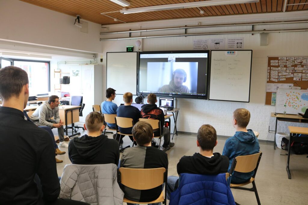 Schüler sitzen in einem Klassenraum und verfolgen eine Videokonferenz.