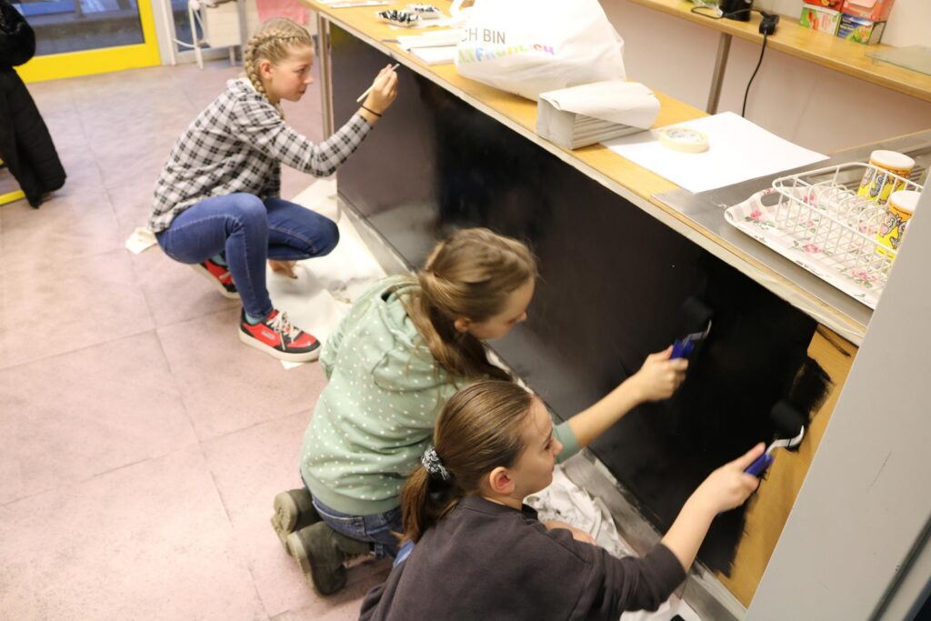 Drei Schülerinnen streichen gemeinsam eine Wand in einem Klassenraum.