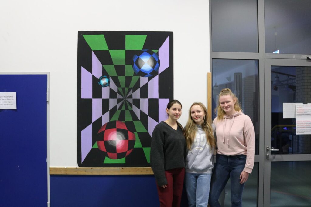 Drei Schülerinnen stehen vor einem großen, bunten Kunstwerk an der Wand.