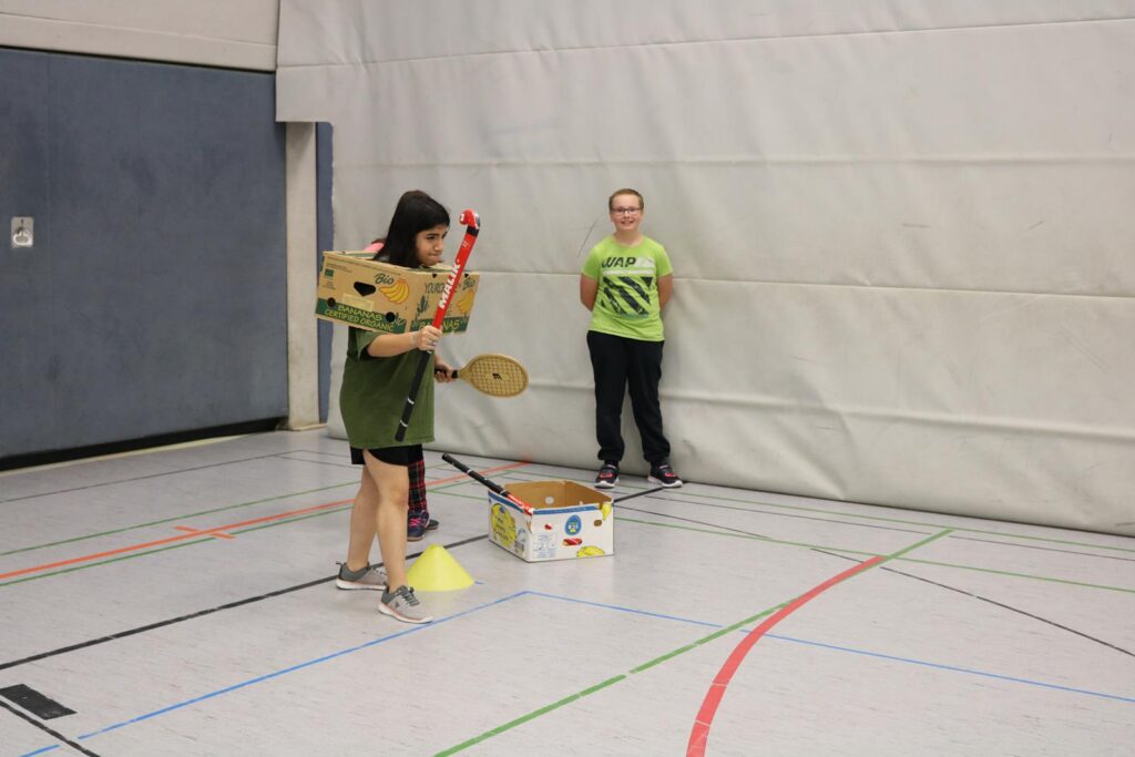 Kinder spielen mit selbstgebastelten Instrumenten in einer Sporthalle.