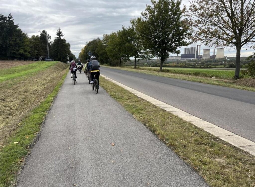 Zwei Radfahrer auf einem Weg neben einer Straße, Bäume im Hintergrund.