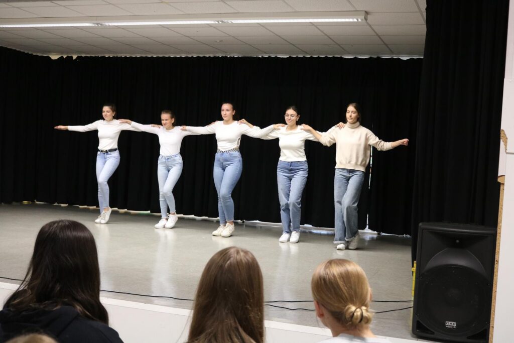 Tanzgruppe auf einer Bühne in einheitlichen Outfits mit Jeans und weißen Oberteilen.