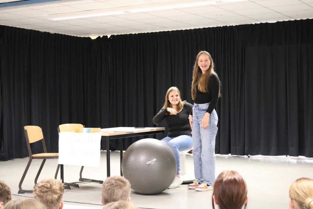Zwei Schülerinnen stehen auf einer Bühne, eine sitzt auf einem Gymnastikball.