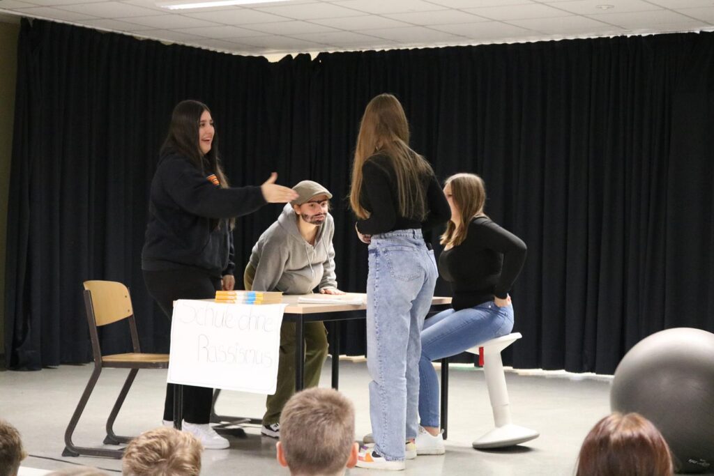 Schülerinnen und Schüler führen eine Szene auf der Bühne vor einer Zuschauergruppe auf.