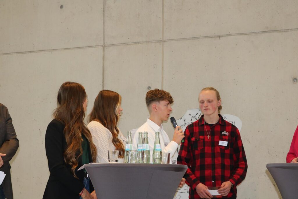 Schüler sprechen auf einer Veranstaltung, Stühle und Tische im Hintergrund.
