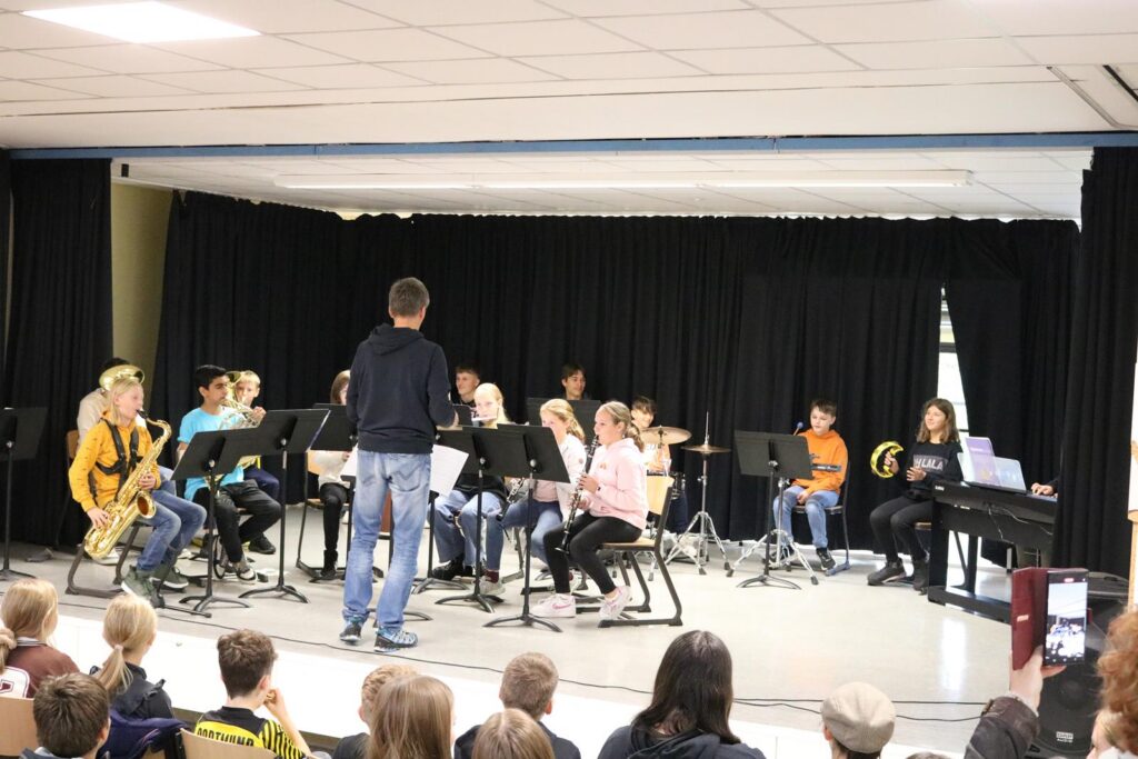Schüler*innen spielen in einem Musikraum vor Publikum. Ein Lehrer leitet das Ensemble.