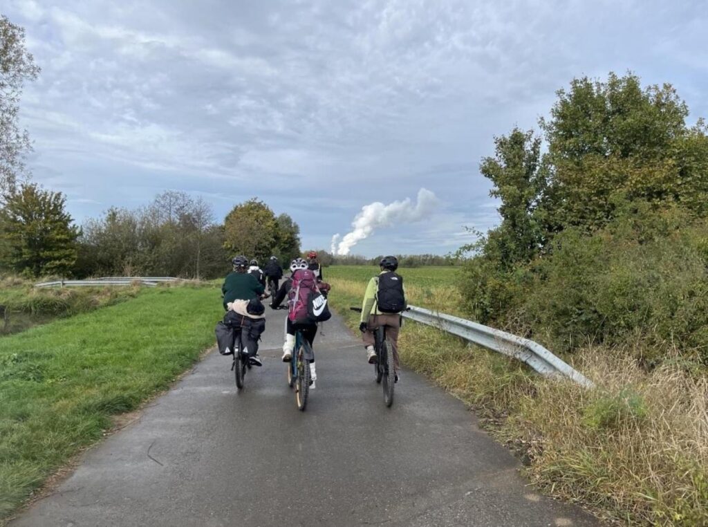 Vier Radfahrer auf einem Weg, umgeben von Bäumen und Wiese, im Hintergrund eine rauchende Fabrik.