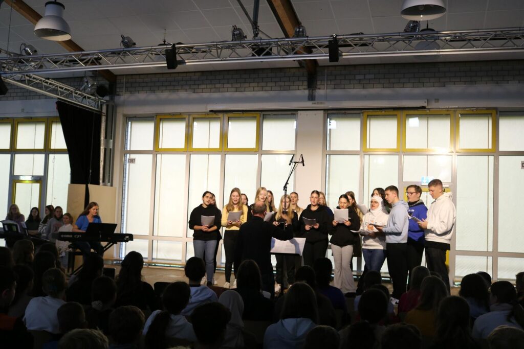 Schüler stehen auf der Bühne und singen. Publikum schaut zu. Ein Klavier ist sichtbar.