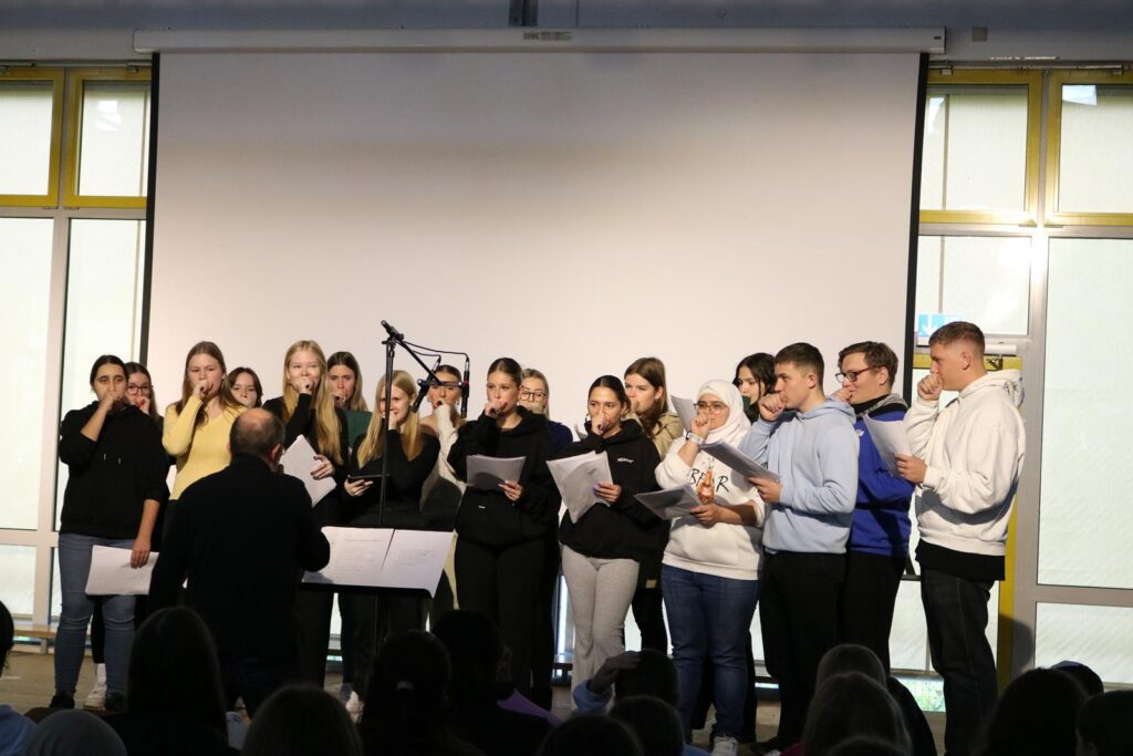Chorgruppe singt auf einer Bühne vor Publikum in einem Klassenraum.