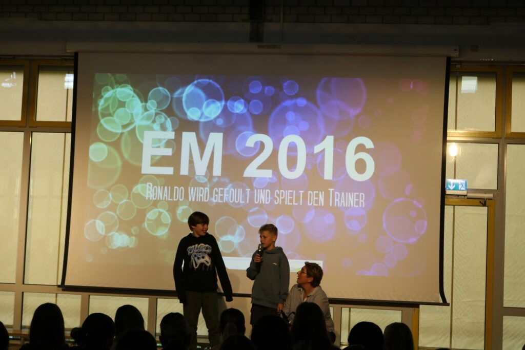Präsentation mit drei Schülern auf der Bühne, Hintergrundbild mit "EM 2016".