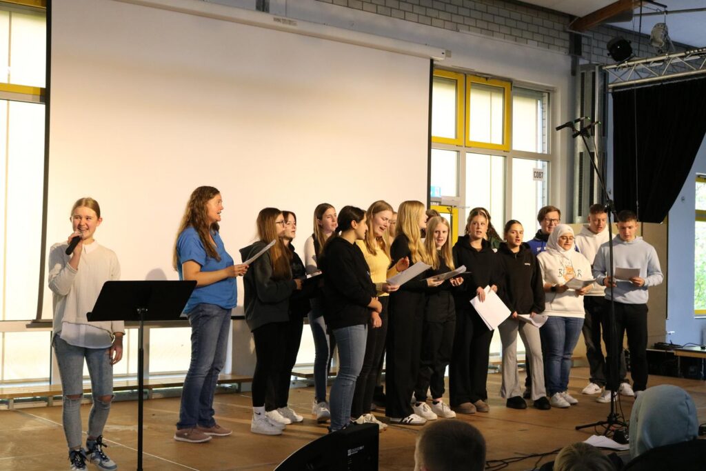Gruppe von Schülerinnen und Schülern singt auf einer Bühne vor Publikum.
