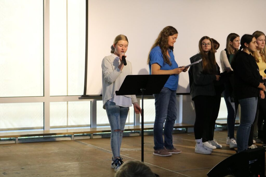Schülerin steht am Mikrofon, während andere Schüler hinter ihr singen oder lesen.
