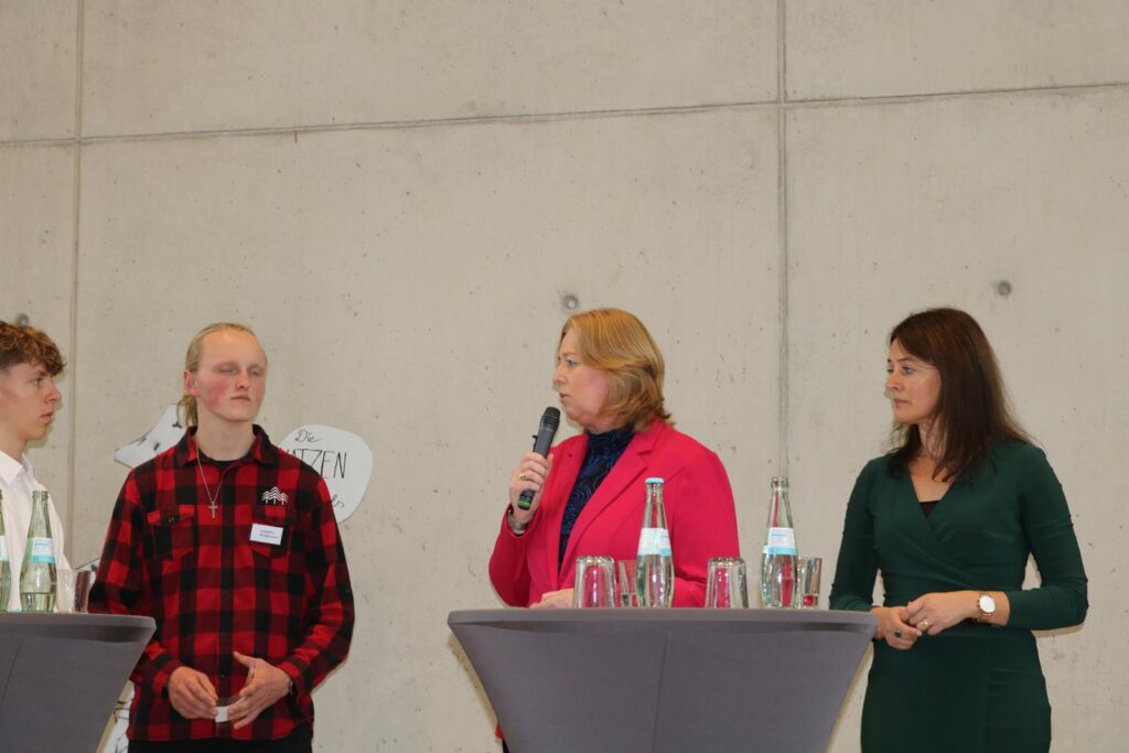 Zwei Personen stehen am Tisch, während eine Frau mit Mikrofon spricht.