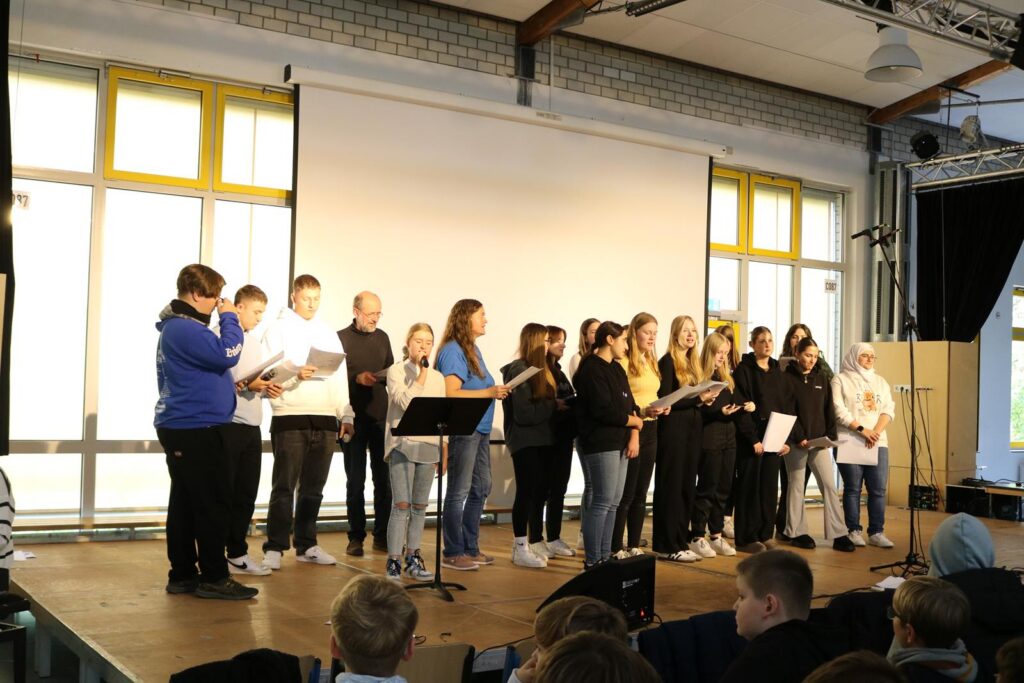 Schülerinnen und Schüler stehen auf der Bühne und präsentieren vor einer Gruppe.