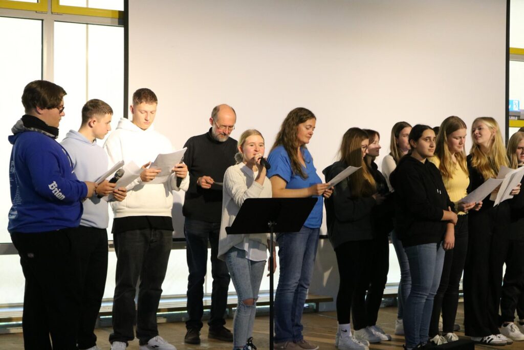 Schüler singen auf einer Bühne, einige halten Notenblätter.