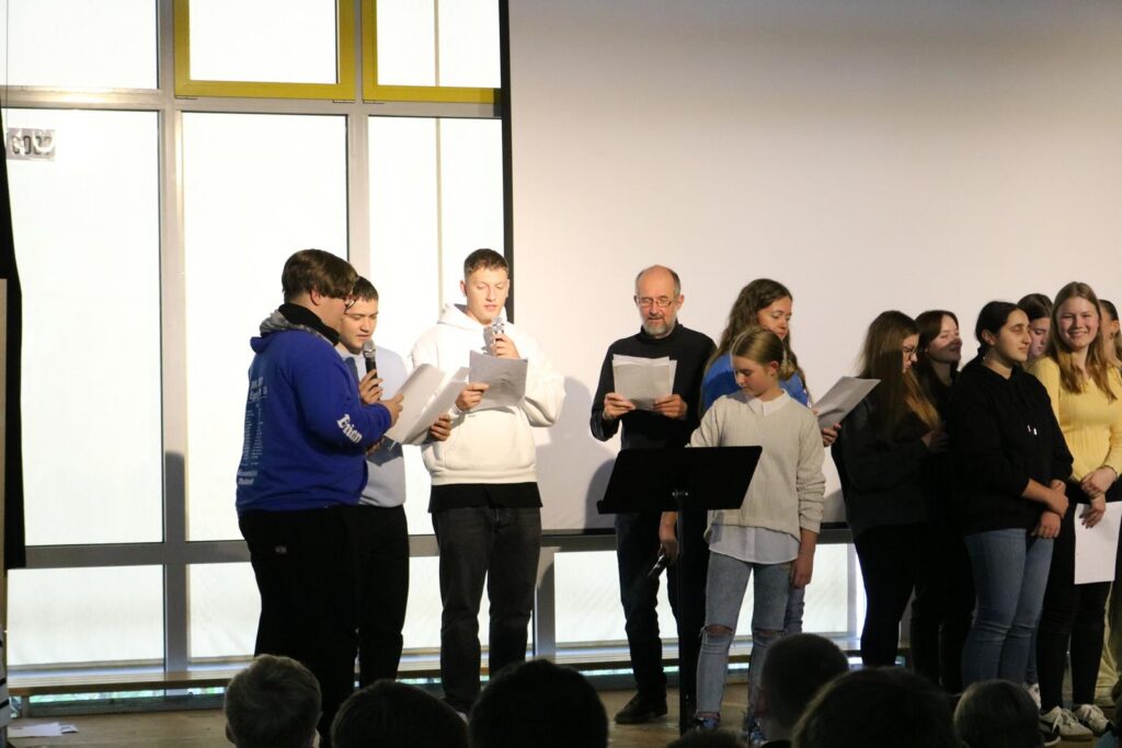 Schülerinnen und Schüler singen auf einer Bühne, einige halten Papier in der Hand.