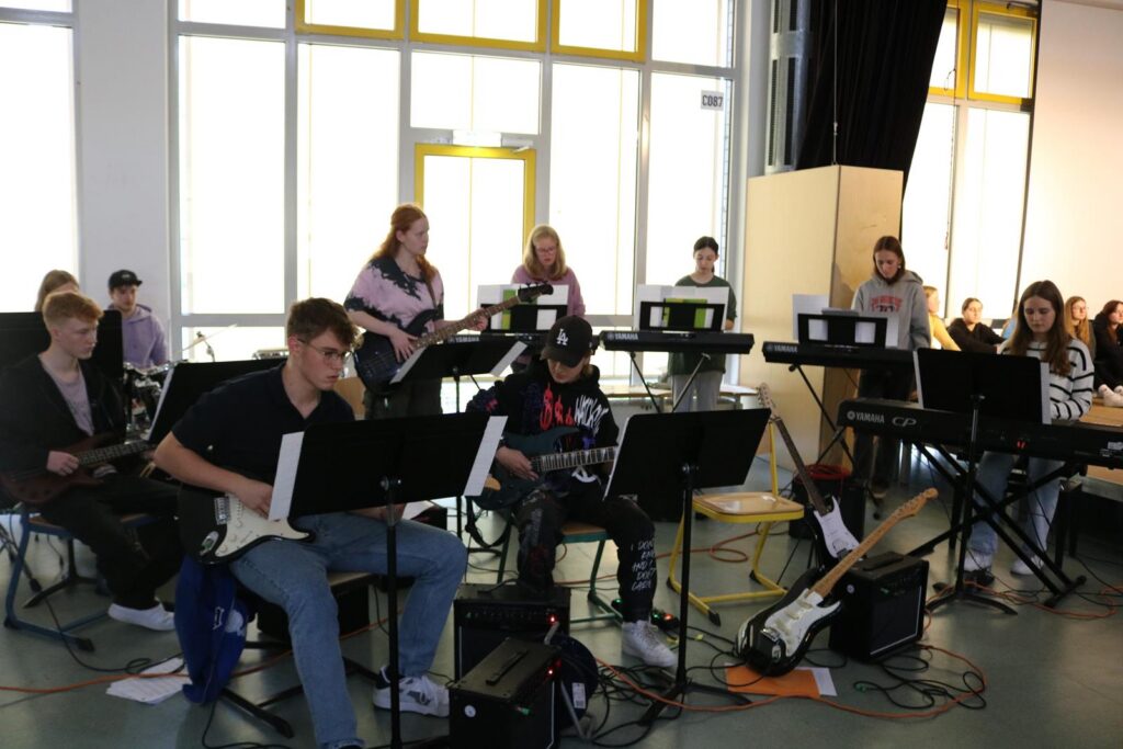 Schüler spielen in einer Aula Musikinstrumente auf Bühnen.