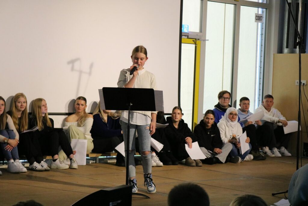 Schülerin steht am Mikrofon vor einer Gruppe sitzender Schüler.