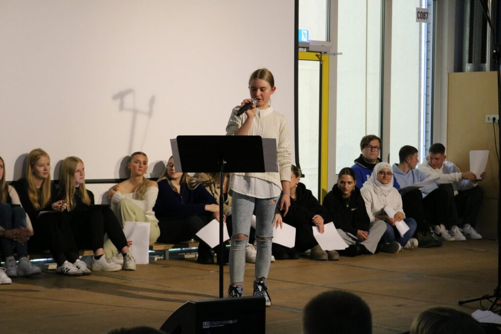 Schülerin steht am Mikrofon, andere Schüler sitzen auf der Bühne.
