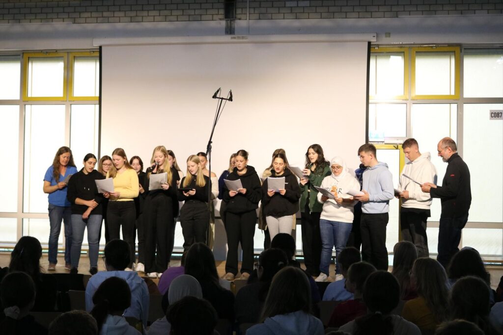 Gruppe von Schülerinnen und Schülern singt auf einer Bühne vor Publikum.