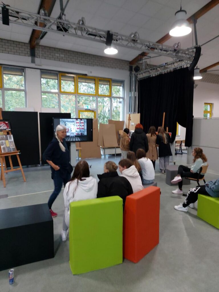 Schüler sitzen auf Sitzmöbeln, während eine Lehrerin erklärt. Im Hintergrund stehen Präsentationswände.