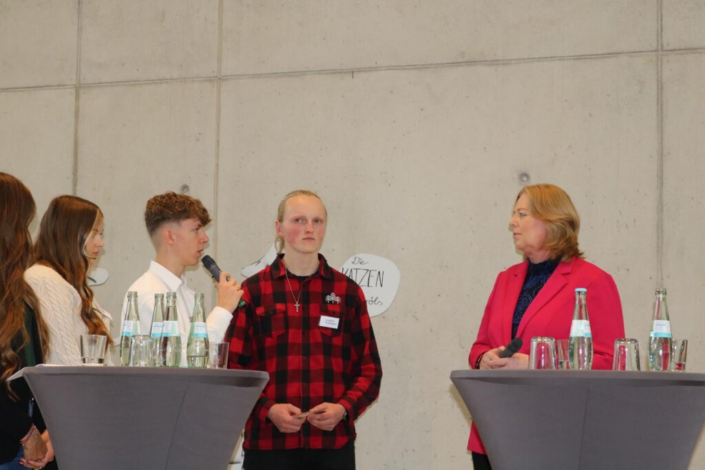 Podiumsdiskussion mit Schülern und einer Rednerin, Getränke auf Tischen.