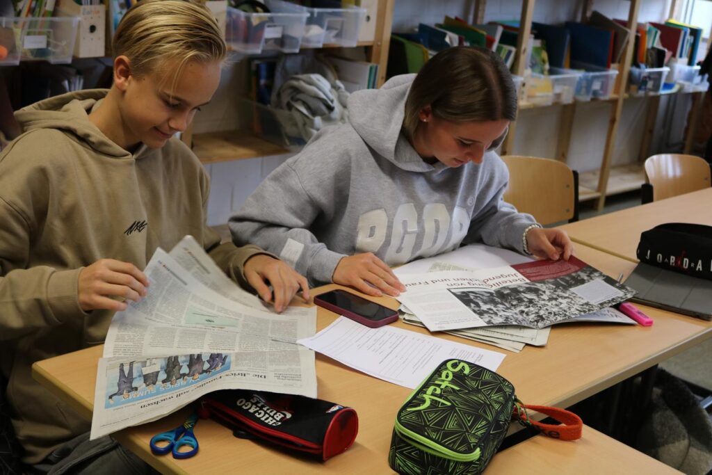 Zwei Schüler arbeiten an einem Tisch mit Zeitungen und Schulmaterialien.
