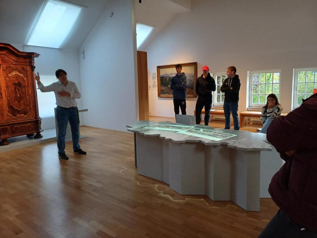 Eine Person erklärt etwas in einem Ausstellungsraum, mehrere Besucher hören zu.