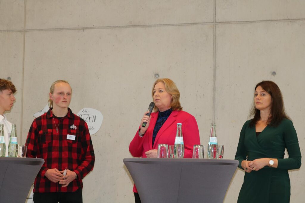 Drei Personen stehen an Stehtischen und sprechen zu einem Publikum.