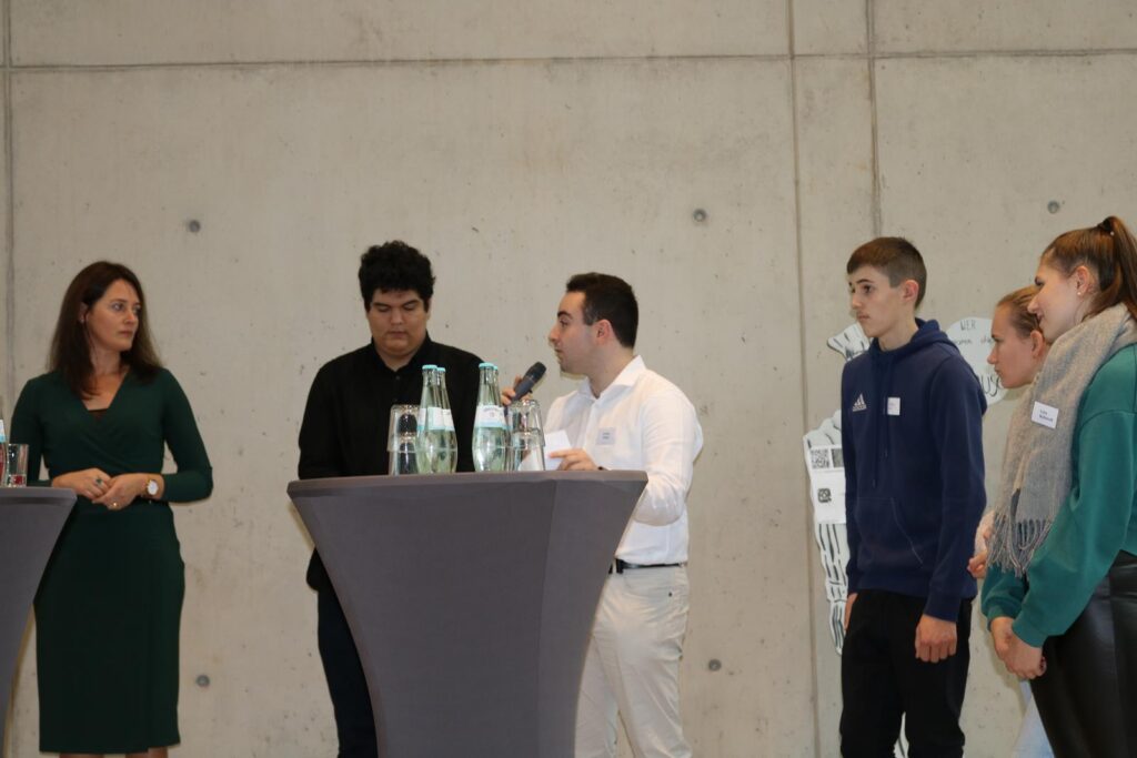 Podiumsdiskussion mit Moderatoren und Schülern an runden Tischen.