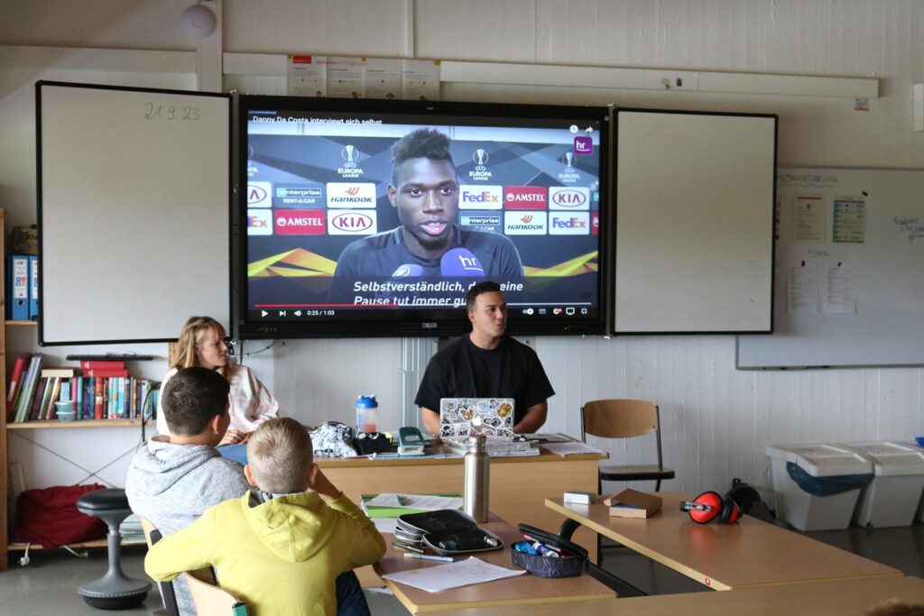 Schüler sitzen an einem Tisch, während eine Präsentation auf einem Bildschirm läuft.