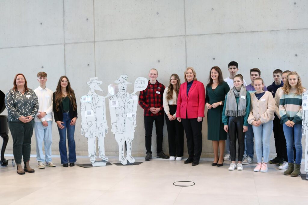 Gruppe von Schülern und Erwachsenen posieren vor grauer Wand mit Pappfiguren.