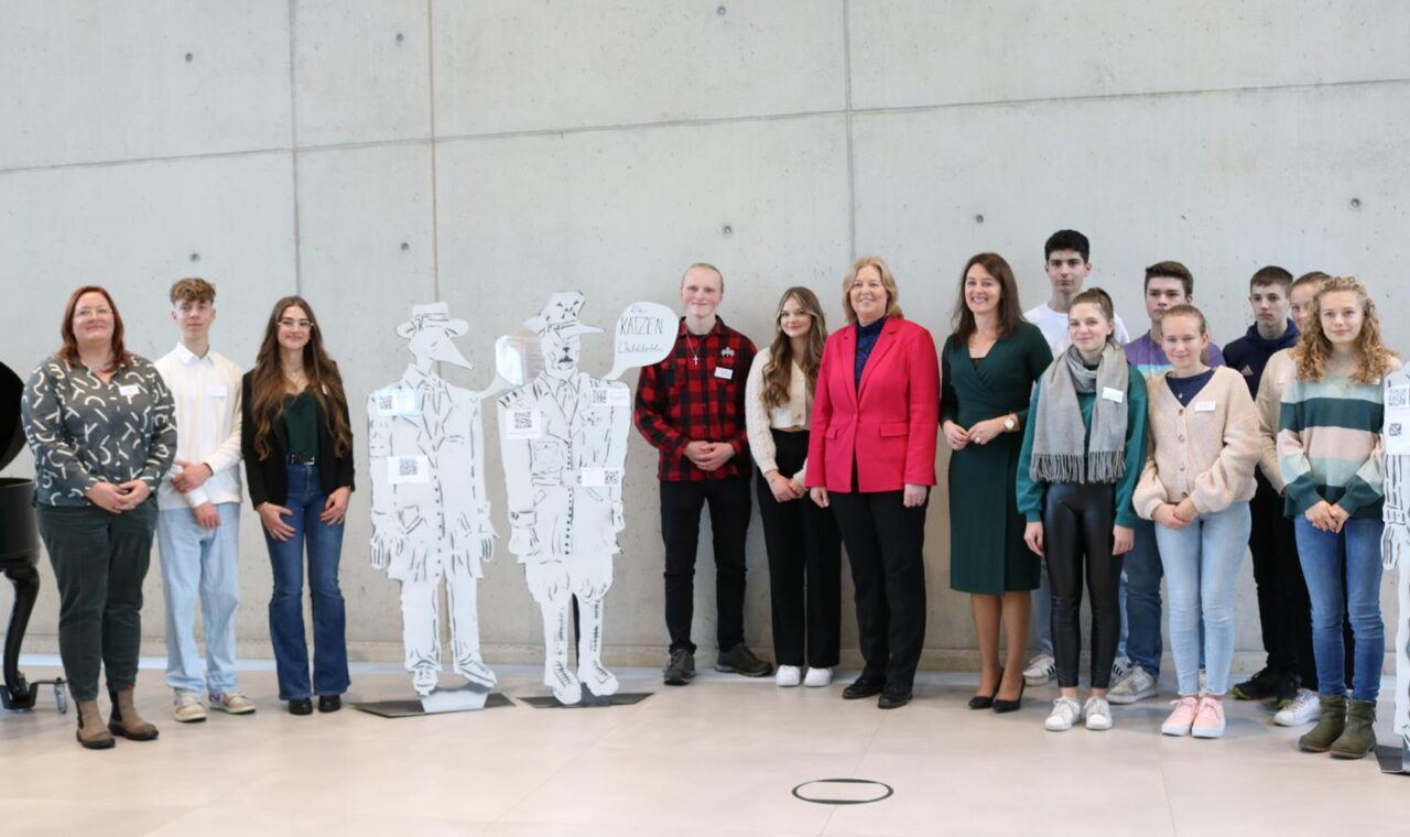 Gruppe von Schülern und Erwachsenen posieren vor grauer Wand mit Pappfiguren.