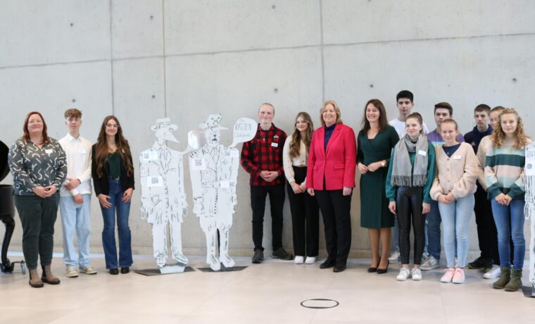 Gruppe von Schülern und Erwachsenen posieren vor grauer Wand mit Pappfiguren.