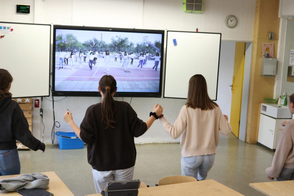 Schülerinnen tanzen vor einem Bildschirm in einem Klassenzimmer.