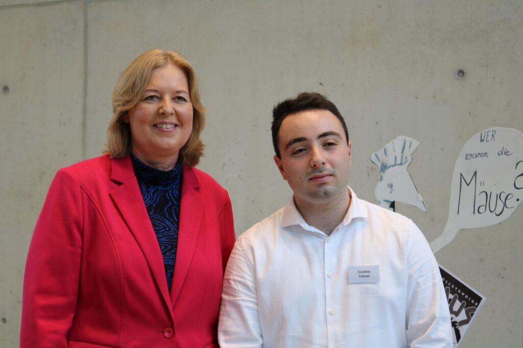 Frau in rotem Blazer und Mann in weißem Hemd stehen vor grauer Wand.