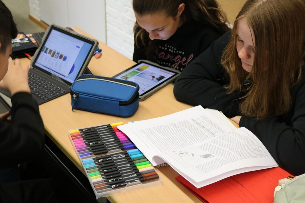 Schüler arbeiten am Tisch mit Tablets und einem Buch, Schreibutensilien liegen bereit.