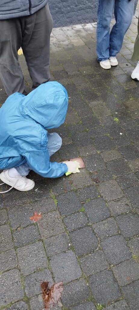 Person in blauer Regenjacke kniet und reinigt den Boden. Umstehende Personen sind zu sehen.