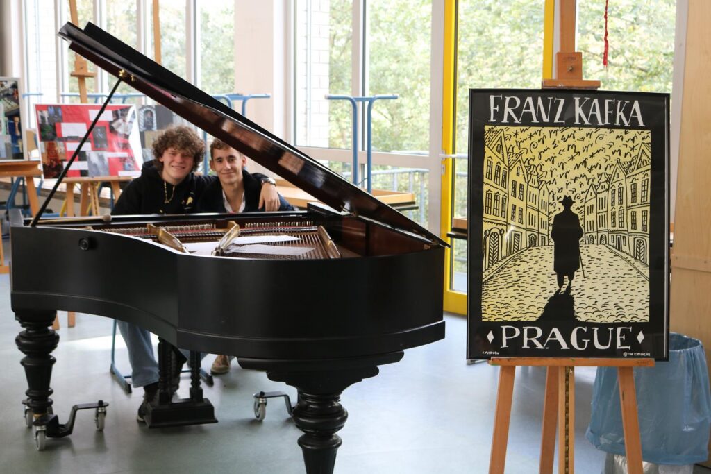 Klavier mit zwei Personen im Hintergrund und Plakat über Franz Kafka in Prag.
