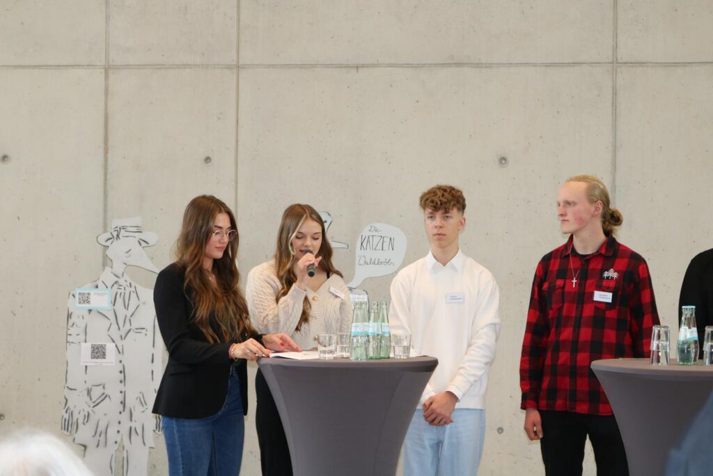 Vier Schüler stehen an einem Tisch und sprechen, im Hintergrund weiße Wand.