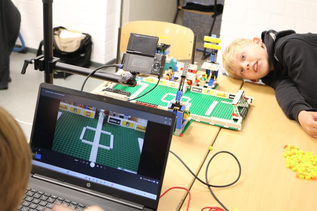 Kinder arbeiten an einem LEGO-Fußballprojekt mit Laptop und Spielfeld.