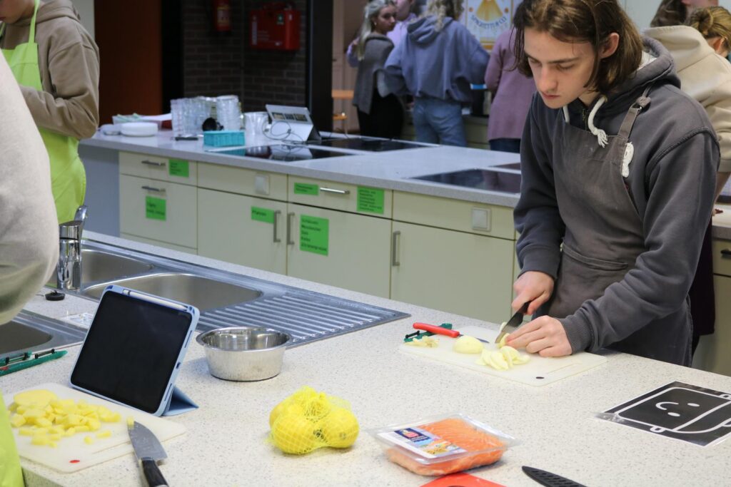 Schüler schneidet Gemüse in einer Schulküche, Tablet auf der Arbeitsfläche.