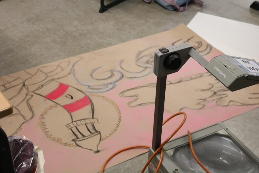 Ein großer, bemalter Karton liegt auf dem Boden, daneben ein Overhead-Projektor.